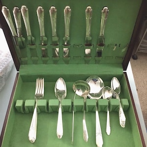 May include: A set of silver-toned flatware arranged in a dark wooden storage case with a bright green felt interior. The collection includes knives, forks, and various spoons, all featuring a polished metallic finish and a classic handle design.
