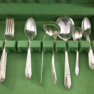 May include: A set of polished silver-toned flatware including a fork, several spoons, and a ladle, arranged in a row. Each piece features a slender handle with a subtle decorative ridge pattern at the base, reflecting light on the metallic surfaces.