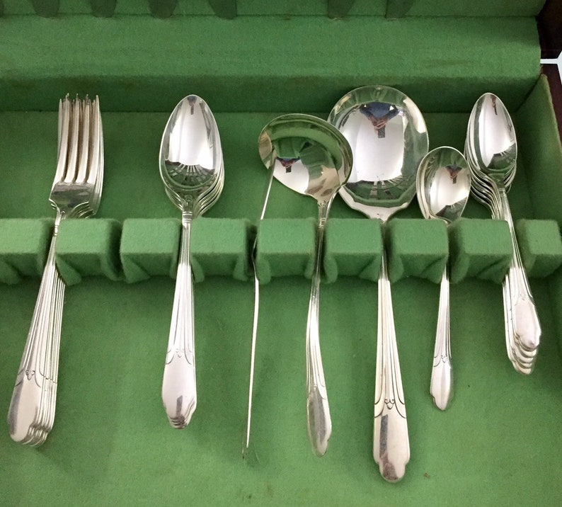May include: A set of polished silver-toned flatware including a fork, several spoons, and a ladle, arranged in a row. Each piece features a slender handle with a subtle decorative ridge pattern at the base, reflecting light on the metallic surfaces.
