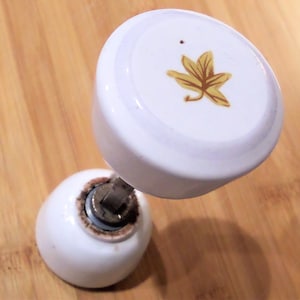 May include: White ceramic cabinet knob with a gold leaf design on top. The knob has a rounded base and a cylindrical top. The hardware is metal. The knob is set against a wooden background.