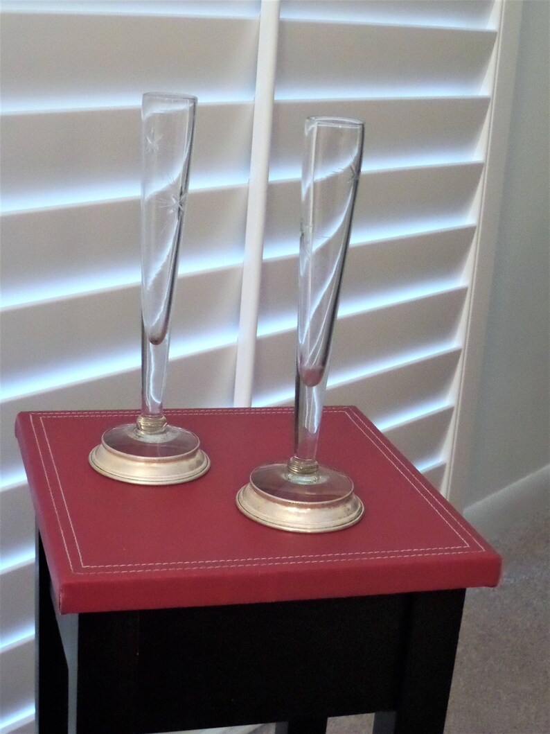May include: Two tall, slender clear glass bud vases with flared openings and circular silver-toned metal bases. Each vase features a delicate, etched starburst pattern on the glass surface, providing a simple and elegant display for single stems.