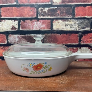 May include: White ceramic casserole dish with a floral design and a glass lid. The dish has a handle on one side.