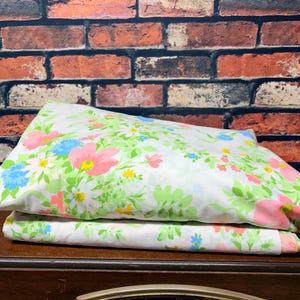 May include: A folded white fabric sheet featuring a repeating floral pattern with large pink blossoms, small blue flowers, and bright yellow centers, accented by light green leaves and stems arranged across the entire surface of the textile.