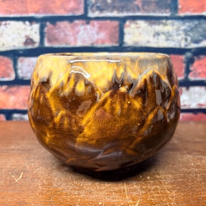 May include: A round ceramic bowl with a glossy finish, featuring a deep brown base color that transitions into a lighter, honey-yellow glaze at the rim. The surface has a textured, organic appearance with visible drips and variations in the glaze.