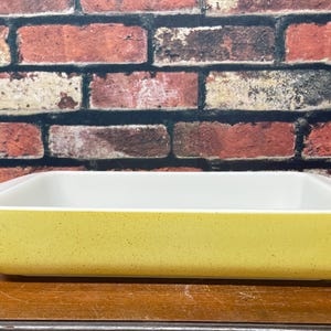 May include: A rectangular baking dish with a yellow speckled exterior and a white interior. The dish is made of ceramic or glass and is sitting on a wooden surface. The background is a brick wall.