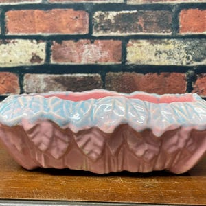 May include: A rectangular ceramic planter featuring a relief pattern of overlapping leaves along the exterior. The base of the planter is a soft pink color, while the upper rim and leaf edges are finished with a glossy, light blue glaze.