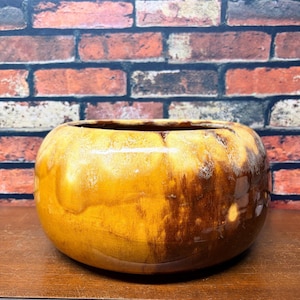 May include: A round ceramic bowl with a glossy finish featuring a blend of warm mustard yellow and deep brown tones. The bowl has a wide, open top and a smooth, curved exterior surface that reflects light, showcasing a mottled, variegated glaze pattern.