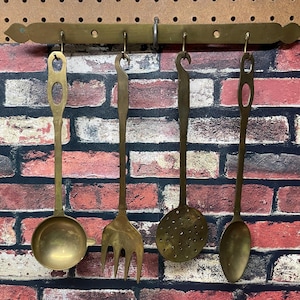 May include: A set of four brass kitchen utensils hanging on a brass rack. The utensils include a ladle, a fork, a slotted spoon, and a spoon. The rack is mounted on a brick wall.