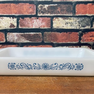 May include: Rectangular, white Pyrex baking dish with a blue floral design. The dish has gently sloping sides and a capacity suitable for casseroles and baked goods. A vintage kitchenware item.