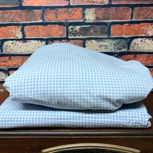May include: Two folded pieces of fabric featuring a light blue and white gingham check pattern. The textile has a small, repeating square grid design throughout, creating a consistent checkered appearance across the stacked layers of material.