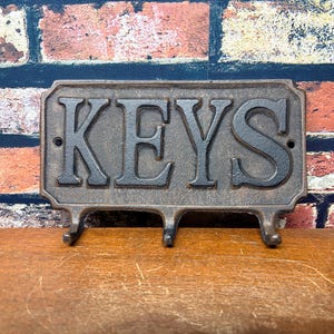 May include: A rectangular dark brown metal wall hook rack featuring the word KEYS in raised, bold, serif lettering. The piece includes three downward-curving hooks at the bottom and two small circular mounting holes on the left and right sides.