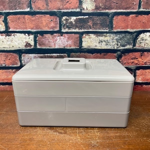 May include: A light gray, three-tiered storage box with a rectangular lid and a small handle. The box is made of a smooth, solid material and sits on a wooden surface against a brick wall background.