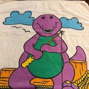 May include: A cream-colored towel featuring a graphic print of a purple dinosaur with a green belly sitting on a wooden dock. The dinosaur holds a yellow fishing rod. Above, there are two blue clouds and two small black birds against a cream background.