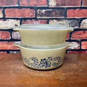 May include: Two vintage Pyrex casserole dishes with clear glass lids. The dishes are beige with a speckled pattern and a blue floral design. They are stacked on top of each other on a wooden surface.