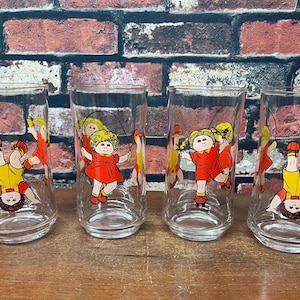 May include: Four clear glass tumblers with Cabbage Patch Kids illustrations. Each glass features a different image of the dolls, including one upside down. The dolls are wearing red and yellow outfits. The glasses are on a wooden surface.