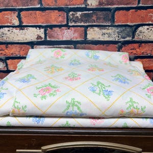 May include: A white fabric sheet featuring a yellow dotted diamond grid pattern with embroidered floral motifs in pink, blue, and green. The textile is folded into layers, displaying a repeating cross-stitch style design across the surface.