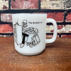 May include: White ceramic coffee mug with a black and white cartoon illustration of a man sitting in a chair watching television. The text "The Breadwinner" is printed on the mug.