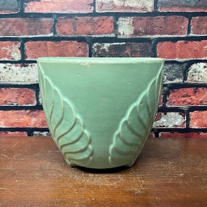 May include: A light green ceramic planter with a leaf design. The planter has a rounded shape and is set on a wooden surface against a brick wall background. The leaf design is a darker shade of green.