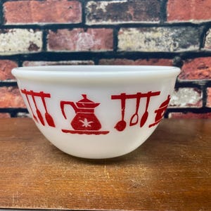 May include: White mixing bowl with red kitchen utensil and teapot designs. The bowl is round and has a wide rim. The red designs are evenly spaced around the bowl. The bowl is sitting on a wooden surface.