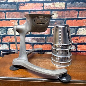 May include: A vintage metal food grater with a hand crank and a black handle. The device features a wide base with suction feet and a hopper for food. Beside the main unit, a stack of four interchangeable stainless steel cylindrical grating drums.