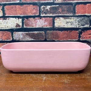 May include: A rectangular, light pink ceramic planter. The planter has rounded corners and a smooth surface. It sits on a wooden surface in front of a brick wall.