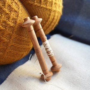 May include: Two wooden spools with white thread wrapped around them. The spools are sitting on a white fabric surface next to a yellow knitted ball.