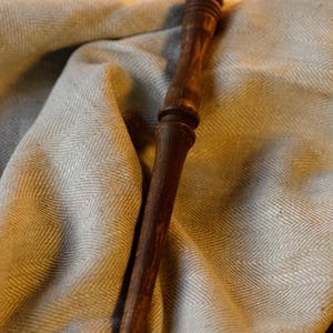 Op de afbeelding: Een donkerbruine houten toverstaf met een afgerond, getextureerd handvat voorzien van horizontale groeven. De staf heeft een gepolijste afwerking en een slanke, langwerpige vorm die taps toeloopt naar een puntig uiteinde.