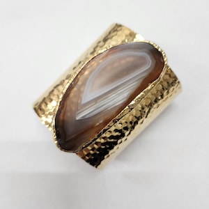 May include: A gold hammered metal cuff bracelet with a brown and white agate stone set into the metal. The stone has a natural, organic shape.