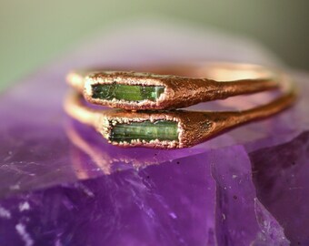 May include: Two copper-colored rings with rectangular green gemstones are stacked on a purple crystal. The rings have a textured, organic design. The gemstones are a vibrant green color, set within the copper bands.