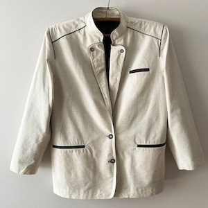 May include: An off-white blazer featuring a mandarin collar, long sleeves, and dark green piping along the edges. The jacket includes two lower welt pockets, one chest pocket, and a two-button front closure, all accented with dark green trim.