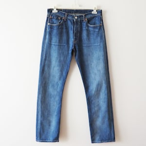 Könnte beinhalten: Blaue Jeans aus Denim mit Knopfleiste und klassischem Fünf-Taschen-Design. Die Jeans hängen an einem weißen Kleiderbügel vor einem weißen Hintergrund. Der Denim hat eine verwaschene Optik mit dezenten Gebrauchsspuren.