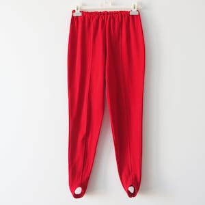 May include: Bright red stirrup pants hanging on a white hanger. The pants have an elastic waistband and a seam down the front of each leg. The stirrups are at the bottom of the legs.