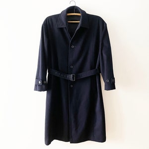 Peut inclure: Un trench-coat bleu marine avec une ceinture assortie et des boutons. Le manteau est suspendu à un cintre en bois sur un fond blanc. Le manteau a des manches longues et un design classique.