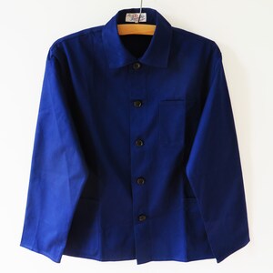 Könnte beinhalten: Eine kobaltblaue Arbeitsjacke mit Knopfleiste, Kragen und einer einzelnen Brusttasche. Die langärmlige Jacke hängt an einem Holzbügel vor weißem Hintergrund. Die Jacke hat vier dunkle Knöpfe.