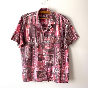 Pode incluir: Uma camisa de manga curta com botões, apresentando um padrão vibrante de formas geométricas, motivos florais e linhas abstratas em vermelho escuro, magenta e branco. A camisa tem um colarinho clássico, fecho frontal e um bolso no peito.