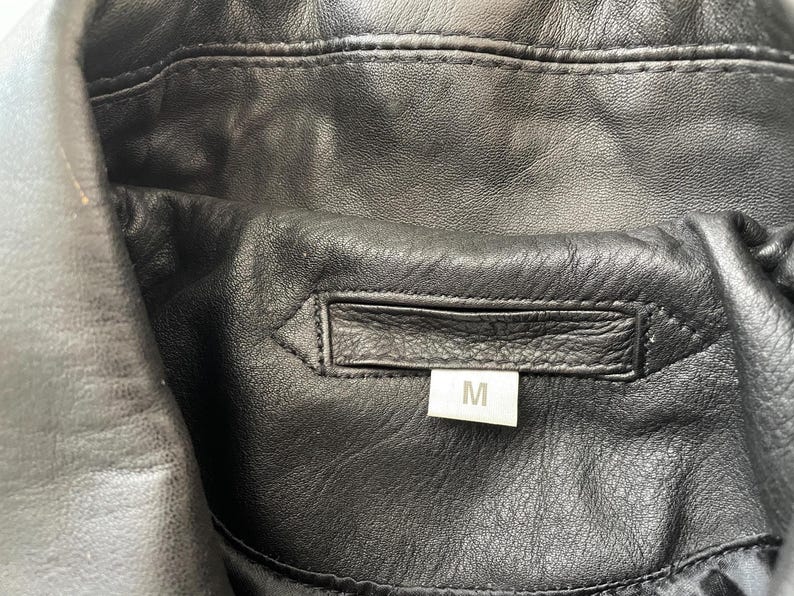 Pu&ograve; includere: Primo piano dell'interno di una giacca di pelle nera. La giacca presenta un colletto cucito e un'etichetta rettangolare con la lettera "M" che indica la taglia. La pelle ha un aspetto strutturato.