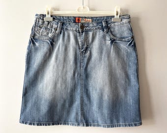 Puede incluir: Una falda vaquera azul claro ligeramente descolorida. La falda tiene una cremallera y un botón de cierre en la parte delantera y dos bolsillos en la parte delantera.