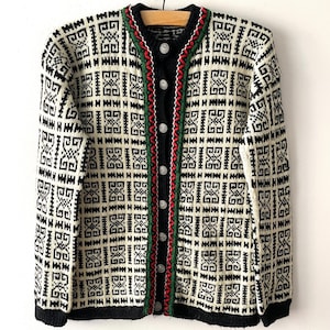 Könnte beinhalten: Eine langärmelige Strickjacke mit einem schwarz-cremefarbenen geometrischen Muster. Das Kleidungsstück hat einen schwarzen gerippten Kragen, Bündchen und Saum sowie eine Knopfleiste vorne. Die Knopfleiste ist mit einer grünen und roten Stickerei verziert.