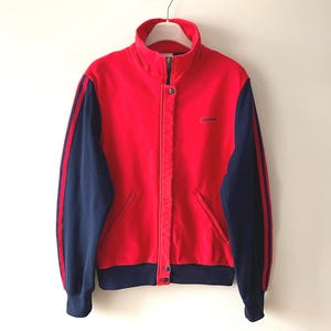 Peut inclure: Veste de survêtement rouge et bleu marine avec col montant et fermeture éclair. Les manches sont bleu marine avec des rayures rouges et un petit logo. Fermeture à boutons et deux poches avant.