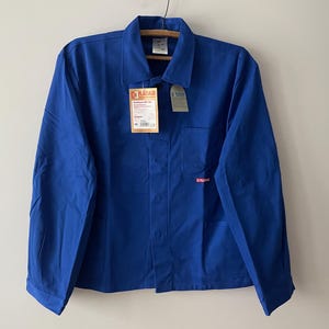 Può includere: Una camicia da lavoro blu brillante con chiusura a bottoni, colletto e taschino sul petto. La camicia ha un'etichetta attaccata con testo. La camicia è appesa a una gruccia di legno.