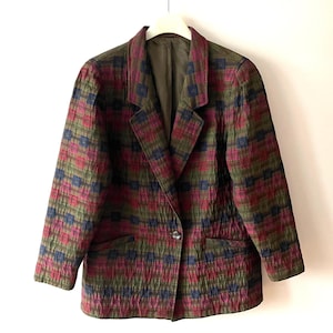 May include: A quilted blazer in a plaid pattern of burgundy, olive green, and navy blue. The jacket has a notched collar, two front pockets, and a single-button closure. The blazer is hanging on a white hanger.