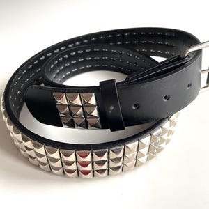 May include: Black leather belt featuring silver pyramid studs. The belt has a silver buckle and a black leather loop. A classic accessory for a punk or rock-inspired style, with a length of approximately 100 cm.