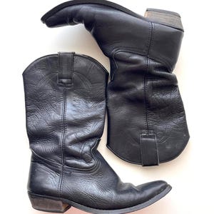Könnte beinhalten: Ein Paar schwarze Cowboystiefel aus Leder mit spitzer Zehenpartie, einem geschichteten Holzabsatz und dekorativen, farblich abgestimmten Nähten am Schaft. Die Stiefel haben Zuglaschen am oberen Rand und eine strukturierte Oberfläche in klassischer Form.