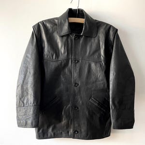 Pu&ograve; includere: Giacca in pelle nera con collo, chiusura con bottoni e due tasche anteriori. La giacca &egrave; appesa a una gruccia di legno su sfondo bianco. Un capo di abbigliamento esterno classico.