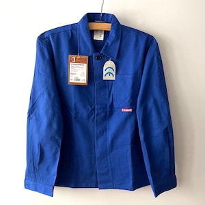Op de afbeelding: Een helderblauwe werkjas met een kraag en knoopsluiting. De jas heeft een borstzak, een klein rood logo en labels met tekst en een CE-markering. Hij hangt aan een houten hanger.
