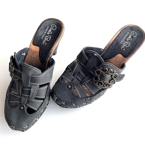May include: A pair of dark gray leather clogs with a wooden sole and a high heel. The clogs feature a cage-like design with decorative metal studs and a buckle closure. The brand name "Carlo Pera" is visible on the insole.