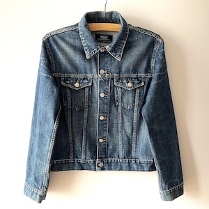 May include: A classic blue denim jacket with a button-up front, two chest pockets, and a collared neckline. The jacket is hanging on a wooden hanger. The denim has a faded appearance.