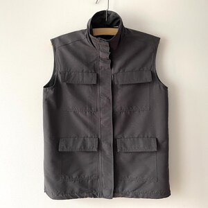 Può includere: Un gilet grigio antracite con collo alto e quattro tasche frontali. Il gilet senza maniche è appeso a una gruccia di legno contro un muro bianco. Il gilet ha un'abbottonatura.