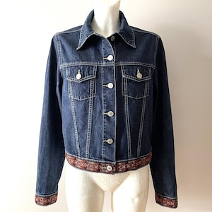 May include: A dark blue denim jacket with a classic collar and button-up front. The jacket features two chest pockets and a decorative patterned band at the hem and cuffs. The jacket is displayed on a mannequin.