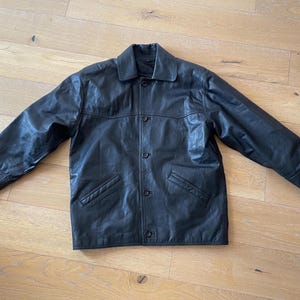 Pu&ograve; includere: Giacca in pelle nera con chiusura a bottoni, due tasche frontali e colletto. La giacca &egrave; stesa su una superficie di legno, mostrando il suo design e la sua consistenza. Le maniche della giacca sono estese.